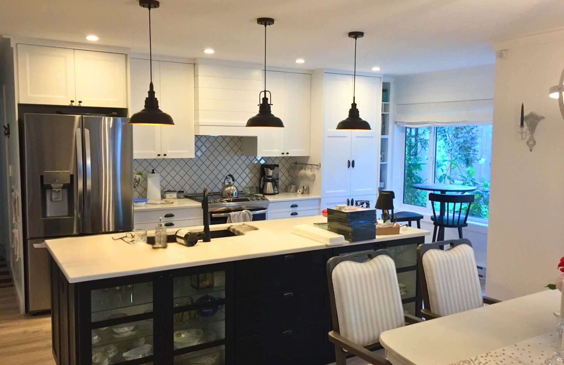 Open white shaker kitchen with a deep navy island, glass-front display cabinets, arabesque tile and three black barn pendants